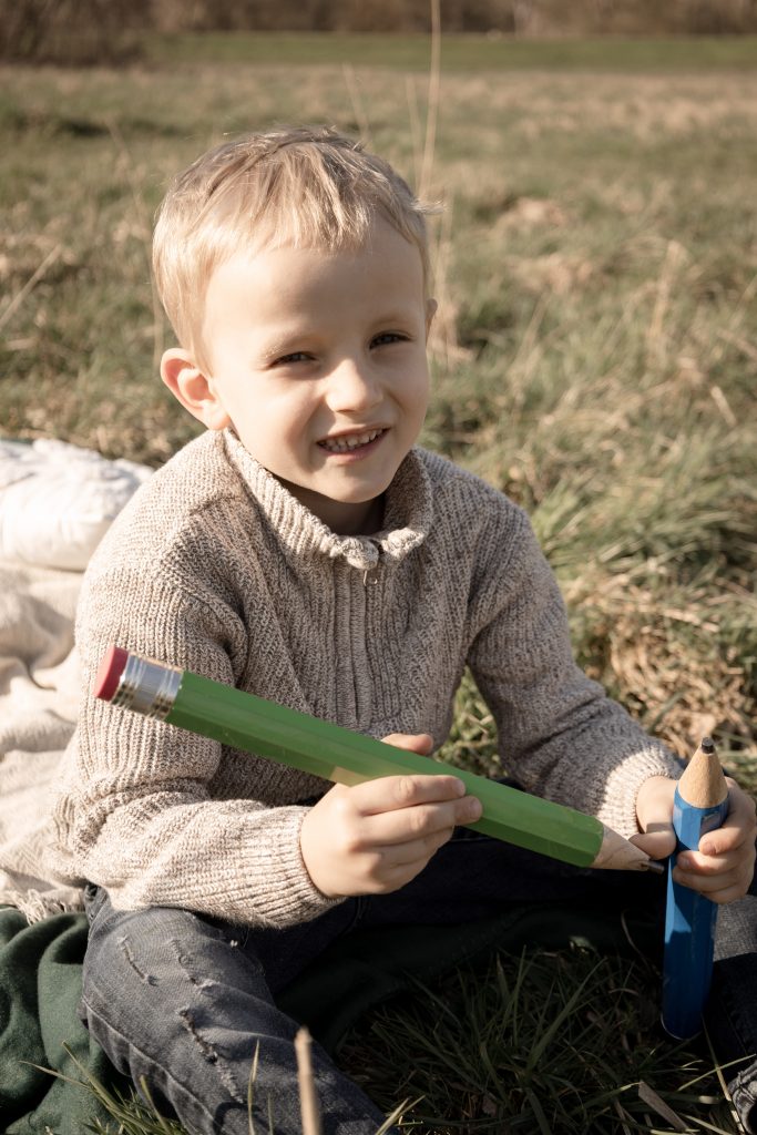 Schulkind sitzt mit Stiften auf einer Decke auf einer Wiese und lacht.