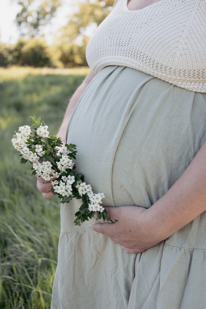 Schwangere hält bei einem entspannten Babybauch-Shooting auf einer Wiese eine Blume in der Hand.