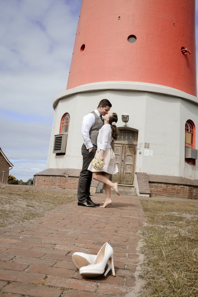 Frisch vermähltes Paar küsst sich innig vor Leuchtturm auf Sylt. Brautschuhe liegen im Vordergrund.